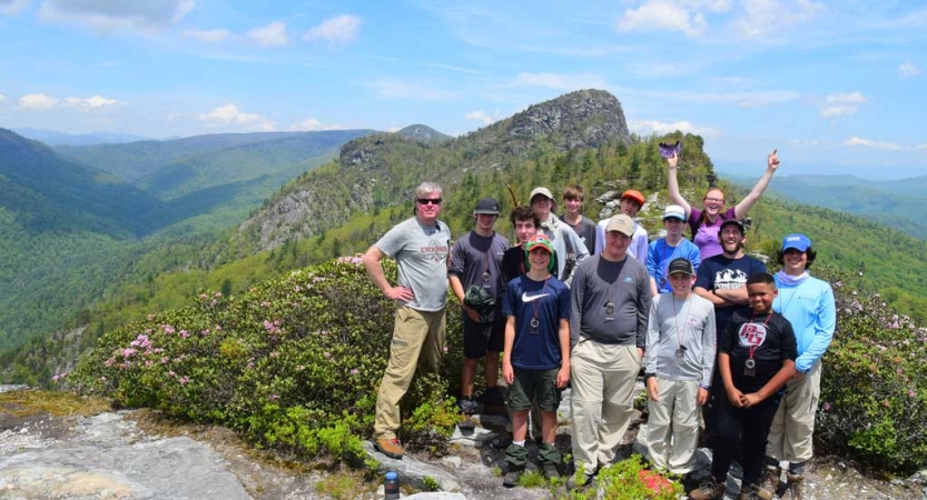 A group of students poses for a photo at high elevation. There is a vast, tree-covered mountainous landscape behind them.